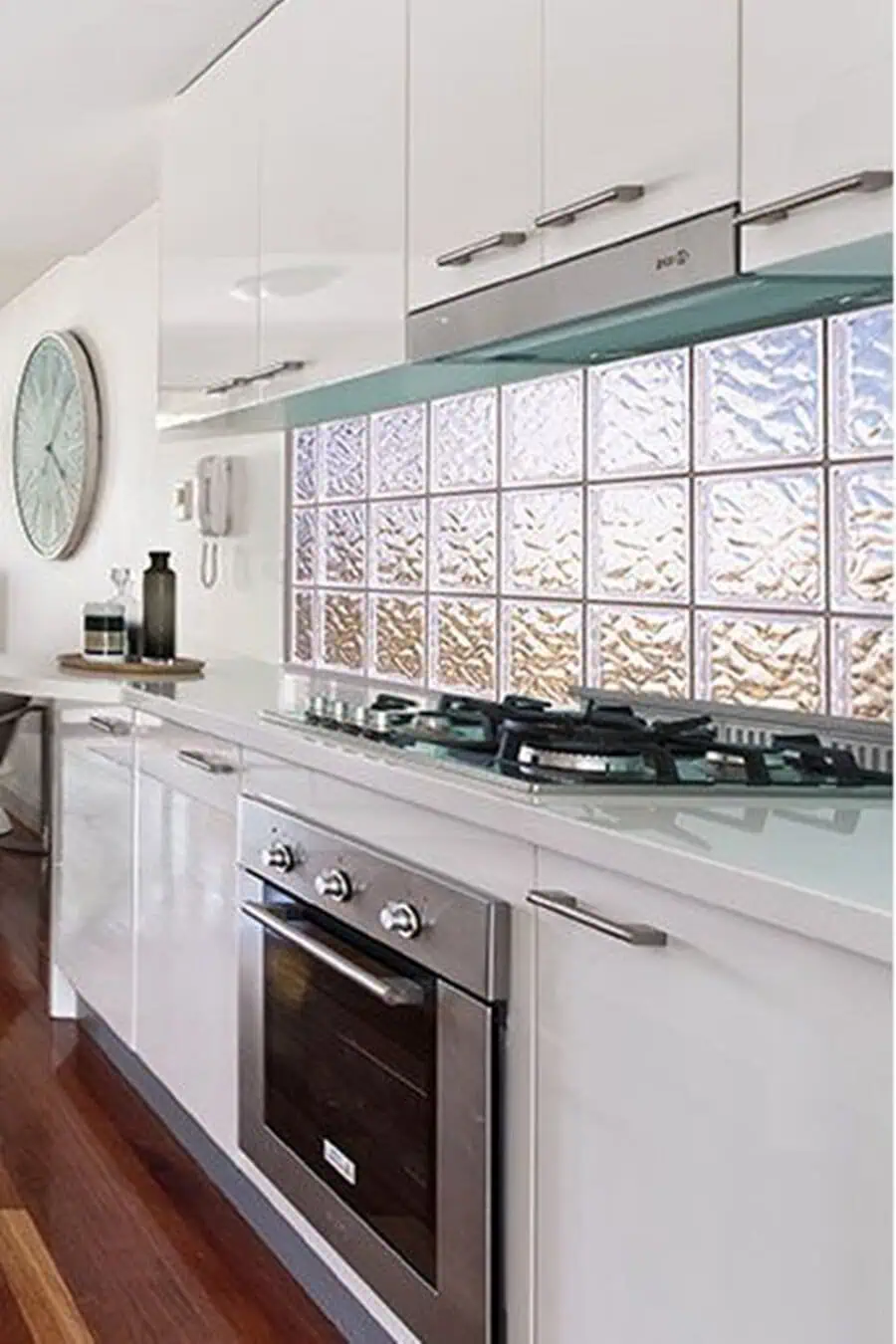 Modern white kitchen with a glass block window backsplash, glossy cabinets, and a polished wood floor.