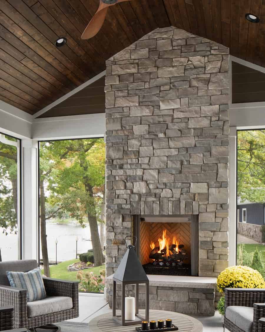 Tall stone fireplace with a bright fire under a dark wood cathedral ceiling, surrounded by windows overlooking a lakeside setting.