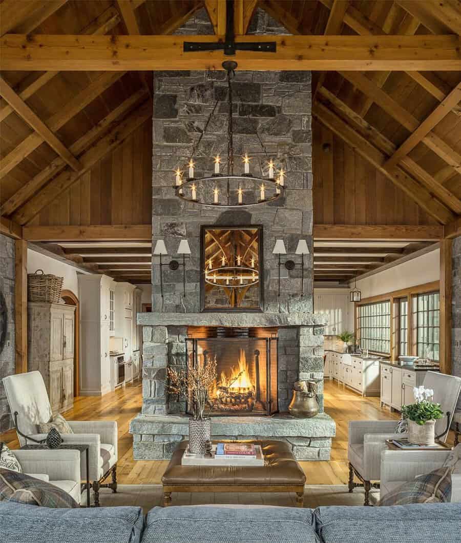 A tall stone fireplace beneath exposed wooden beams and a circular chandelier, set in a warm, rustic living room.