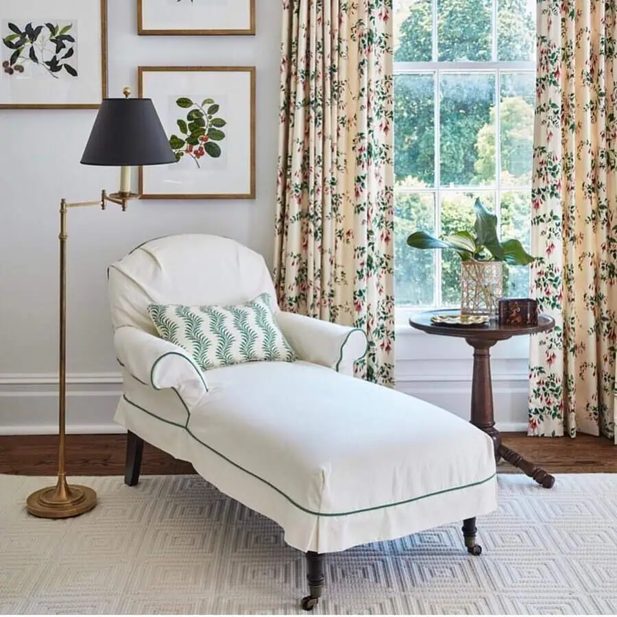 Grandmillennial-style bedroom sitting area with a slipcovered chaise lounge, floral curtains, botanical artwork, and vintage-inspired decor.