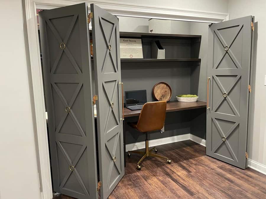 Closet office with gray paneled doors, floating shelves, a dark wood desk, and a brown leather chair in a modern neutral space.