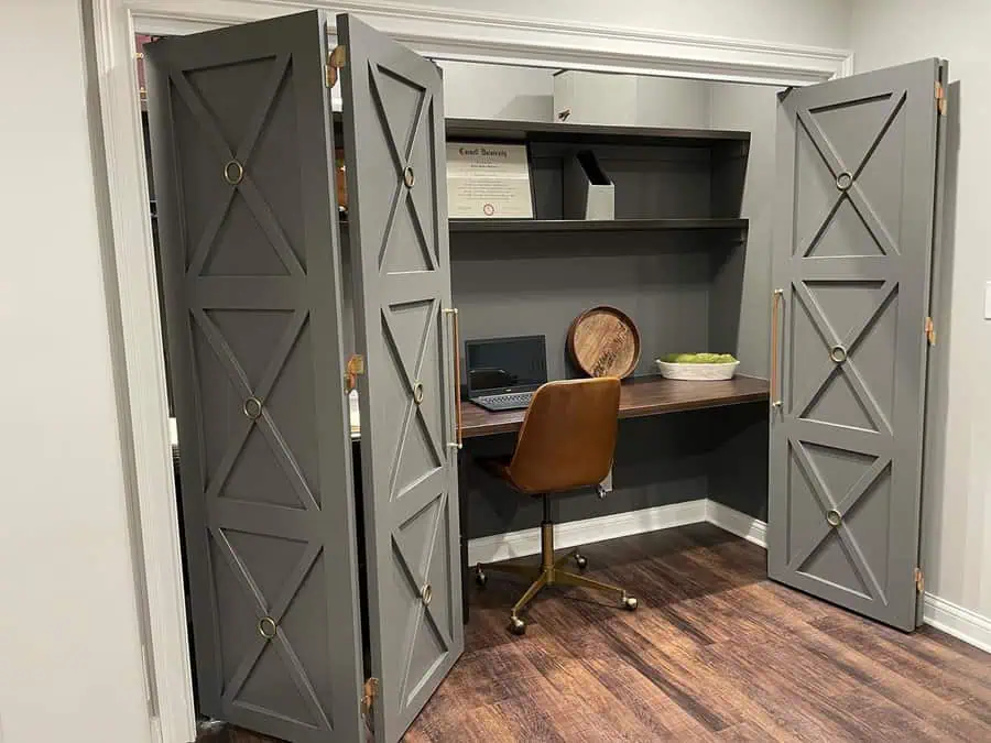 Closet office with gray paneled doors, floating shelves, a dark wood desk, and a brown leather chair in a modern neutral space.