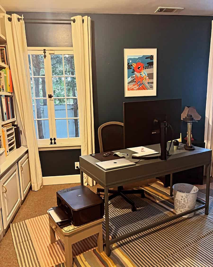 Home office with dark blue walls, a gray desk, and striped rug, featuring light curtains and colorful artwork for balance.