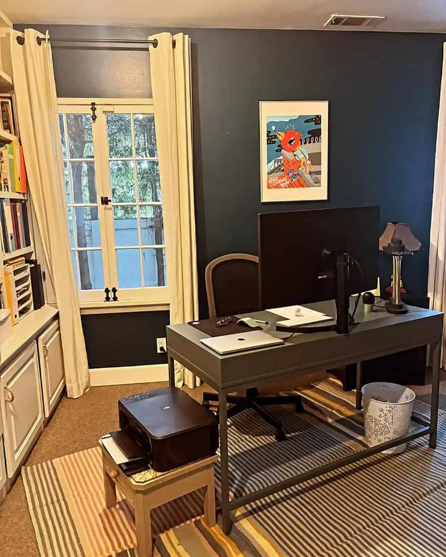 Home office with dark blue walls, a gray desk, and striped rug, featuring light curtains and colorful artwork for balance.