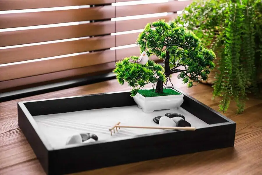 Tabletop zen garden featuring white sand, a bonsai tree with a small bird figurine, black stones, and a rake for a calming setup.
