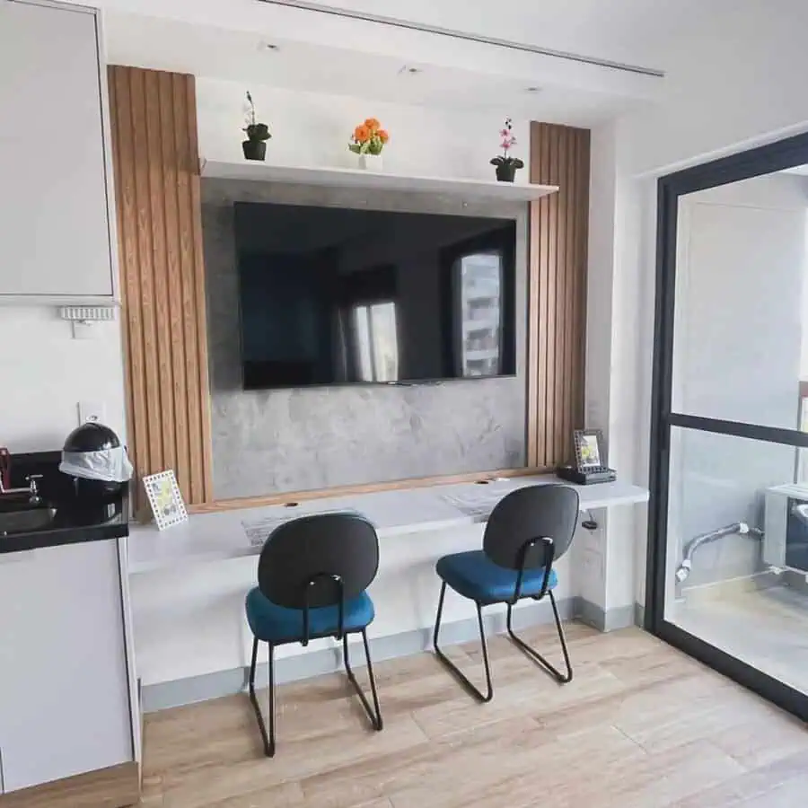 Dual-purpose nook featuring a large wall-mounted TV, slim desk, two blue chairs, and small potted plants overhead in a bright, modern space.
