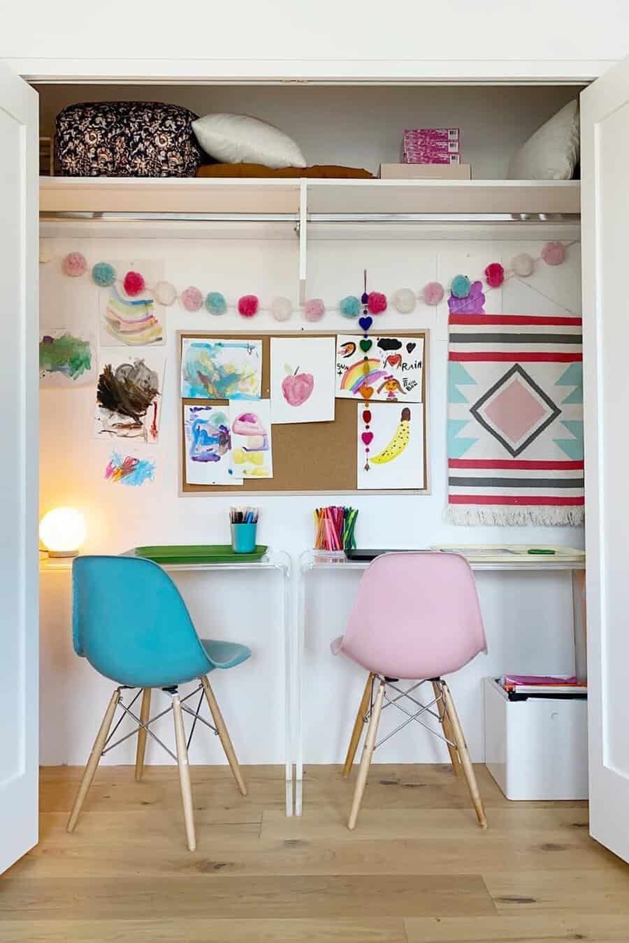 Closet workspace for kids with clear desks, pastel chairs, a corkboard for artwork, a pom-pom garland, and storage on the top shelf.