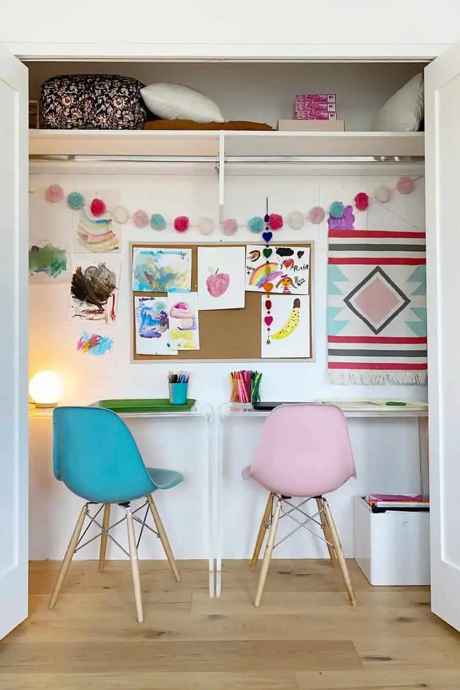 Closet workspace for kids with clear desks, pastel chairs, a corkboard for artwork, a pom-pom garland, and storage on the top shelf.