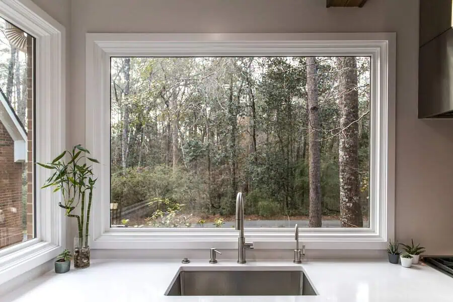Kitchen with a wide picture window overlooking a wooded backyard, white counter, and stainless faucet.