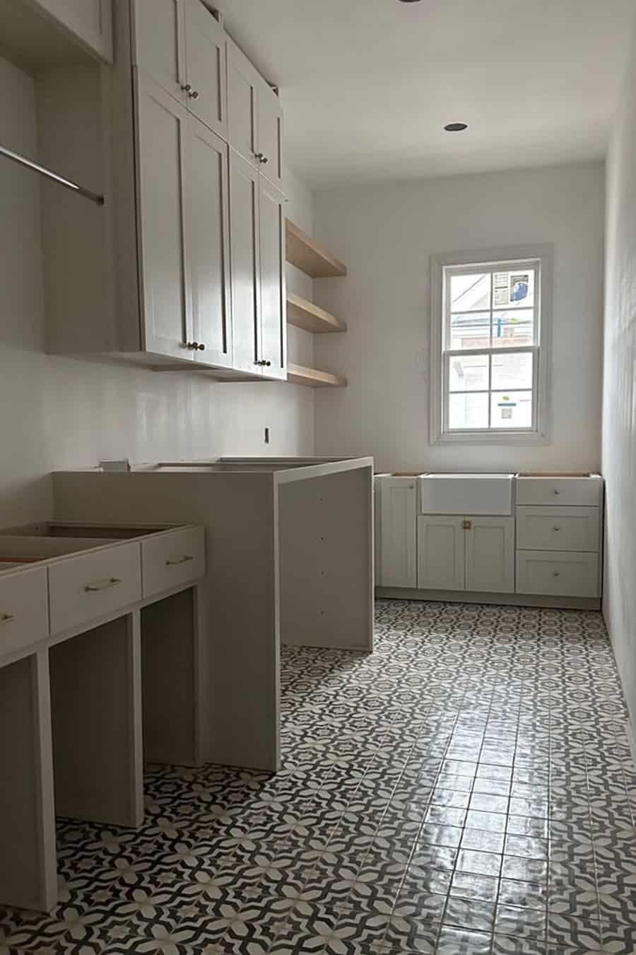 Long laundry room with greige cabinets, open shelves, and a patterned tile floor under a small window.