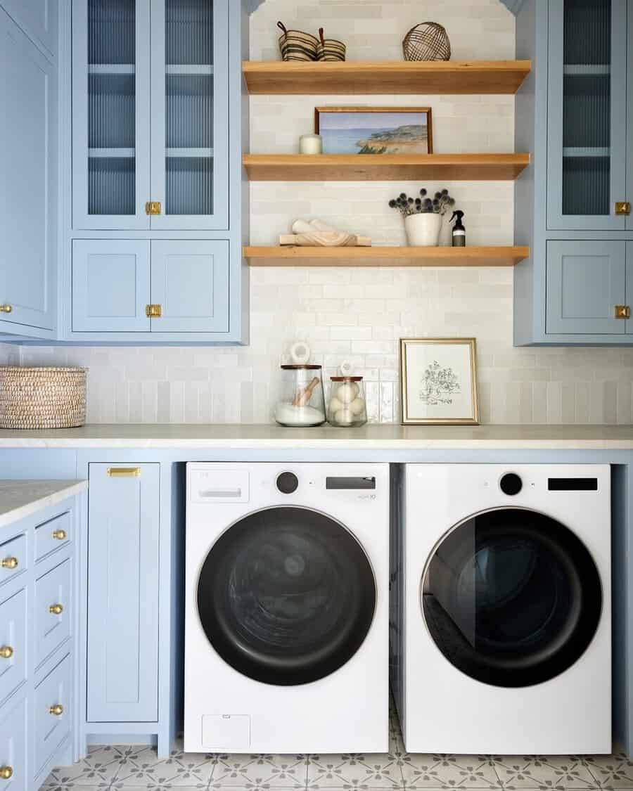 Laundry room with light blue cabinets, wood shelves, brass hardware, white tile backsplash, and patterned floor tile.