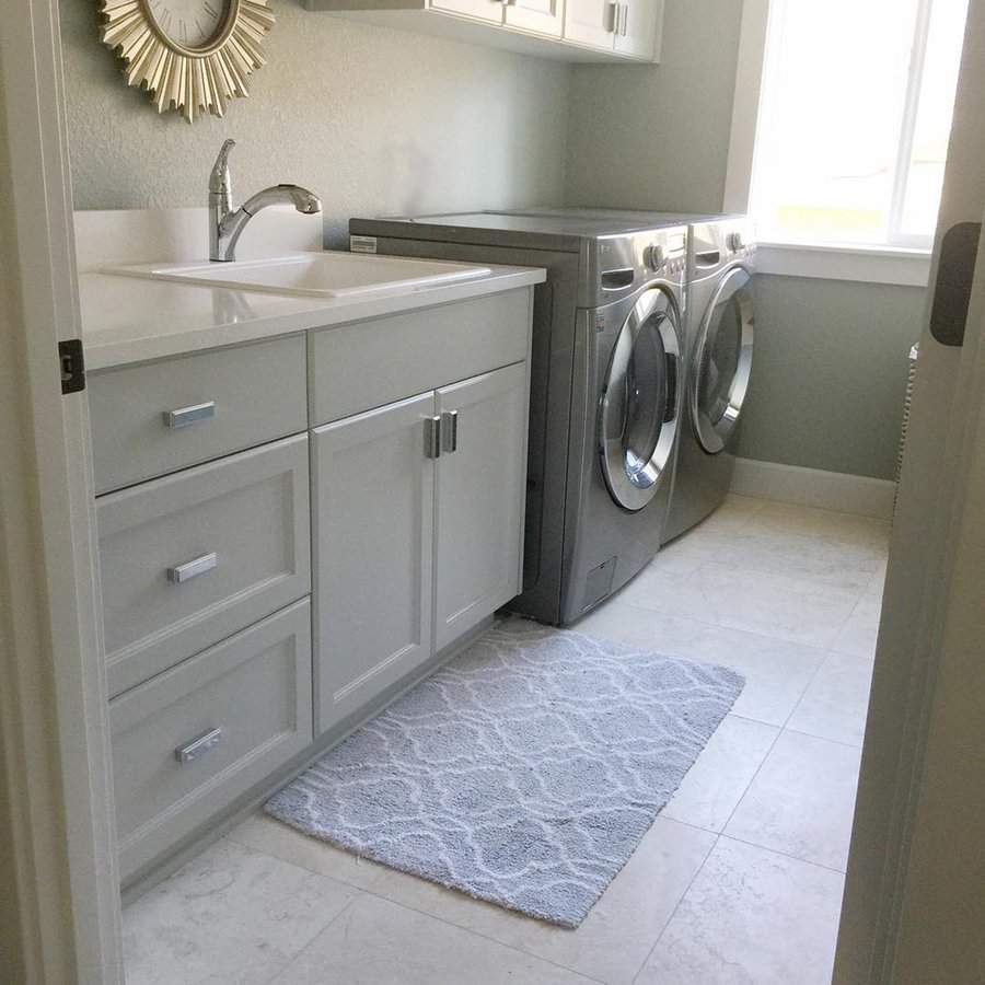 Gray laundry room with front-loading washer and dryer, sink area, and light tile flooring near a sunny window.