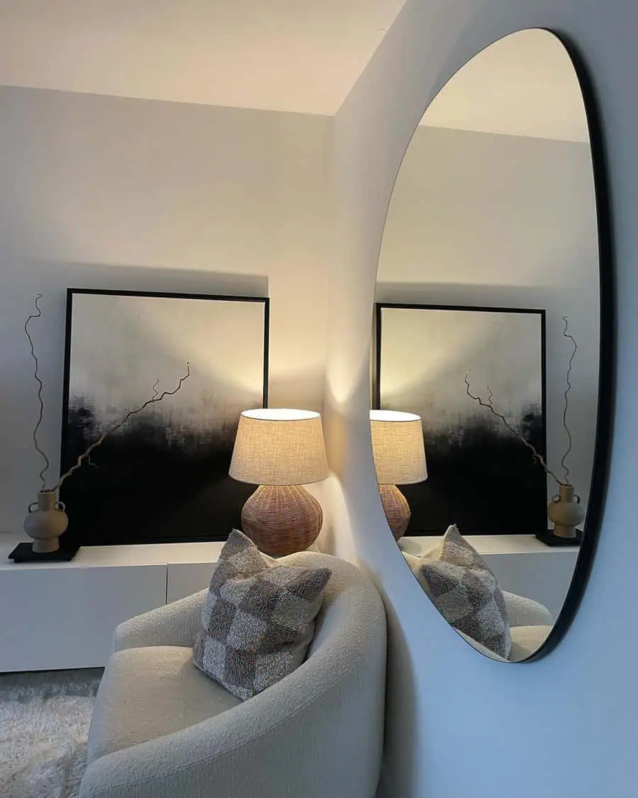 Curved mirror reflecting abstract black-and-white art, a warm table lamp, and a rounded chair in a modern, cozy corner.