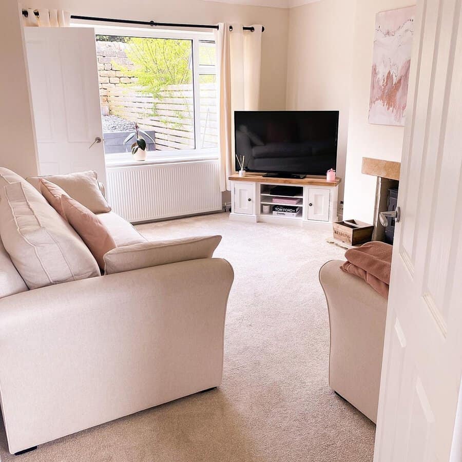 Bright living room with cream sofa, pastel cushions, a TV console, and a large window revealing a garden wall.