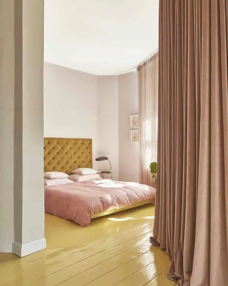 Bedroom with a yellow painted floor, pink bedding, tufted mustard headboard, and warm drapes.