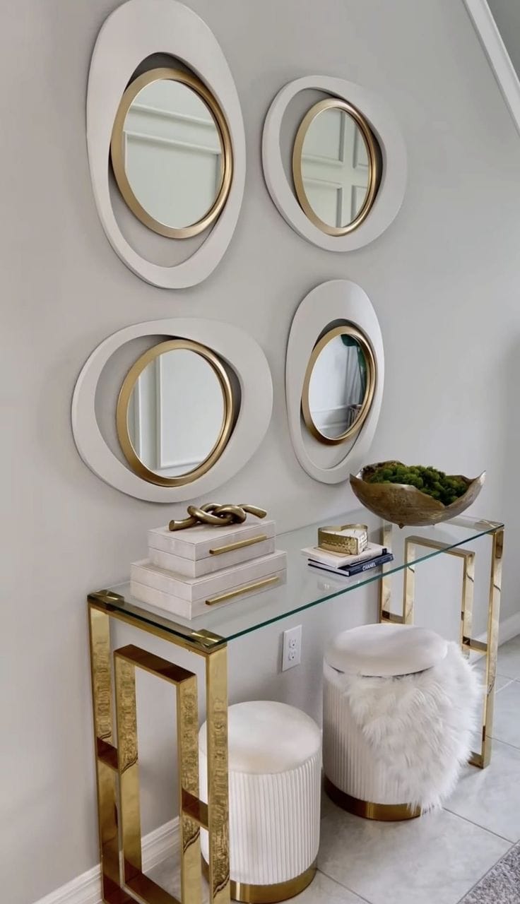 Group of circular mirrors with gold frames above a modern console table with decorative accessories and plush stools.