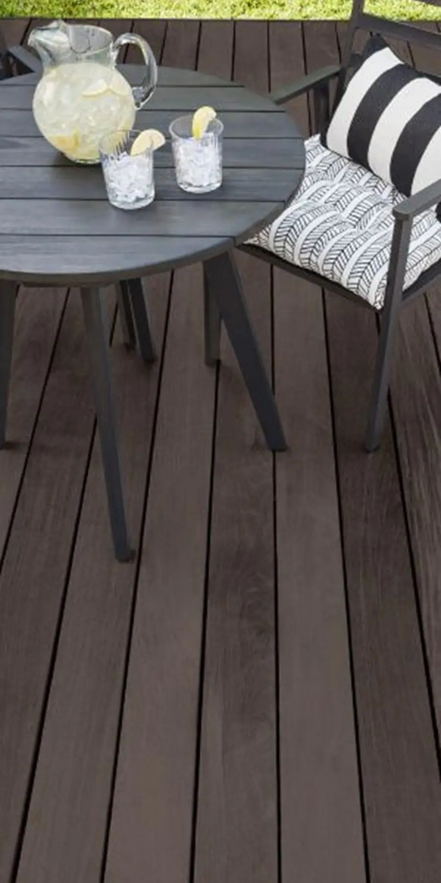 Dark-stained wood deck with a black round table, striped cushion chair, and a pitcher of lemonade.
