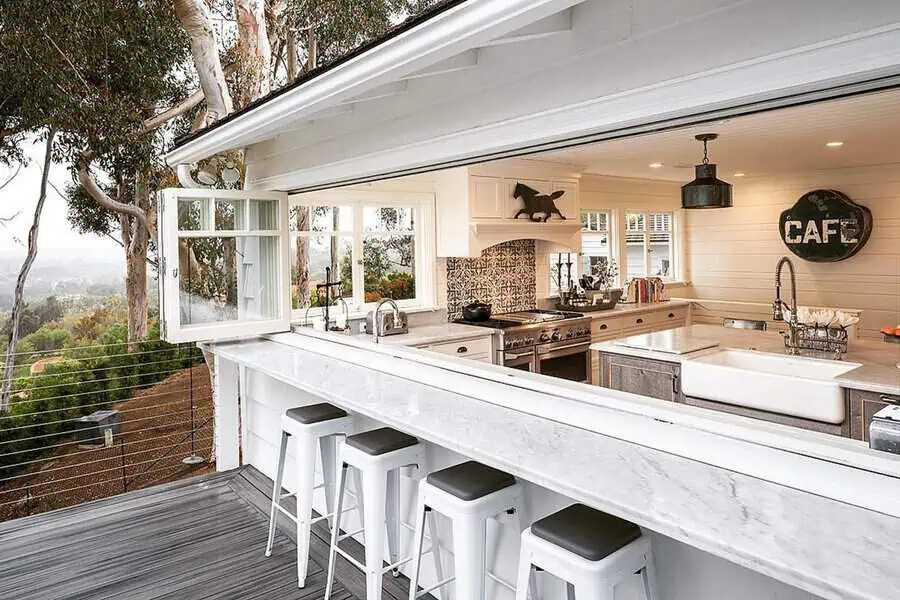Kitchen pass-through window with a marble counter, white stools, and shiplap walls, overlooking a scenic outdoor space.