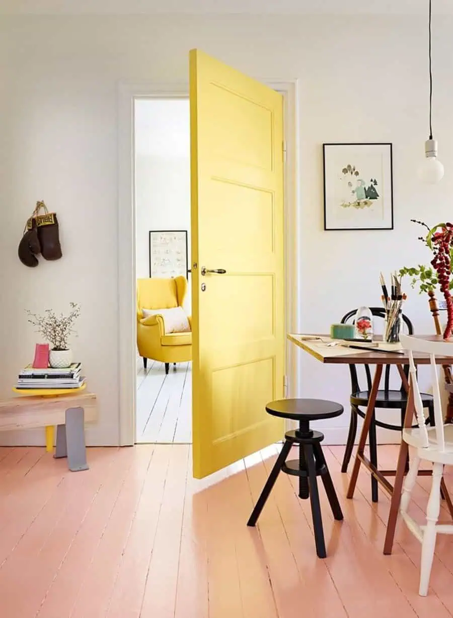 Room with a pink painted wood floor, bright yellow door, and simple wood furniture against neutral walls.