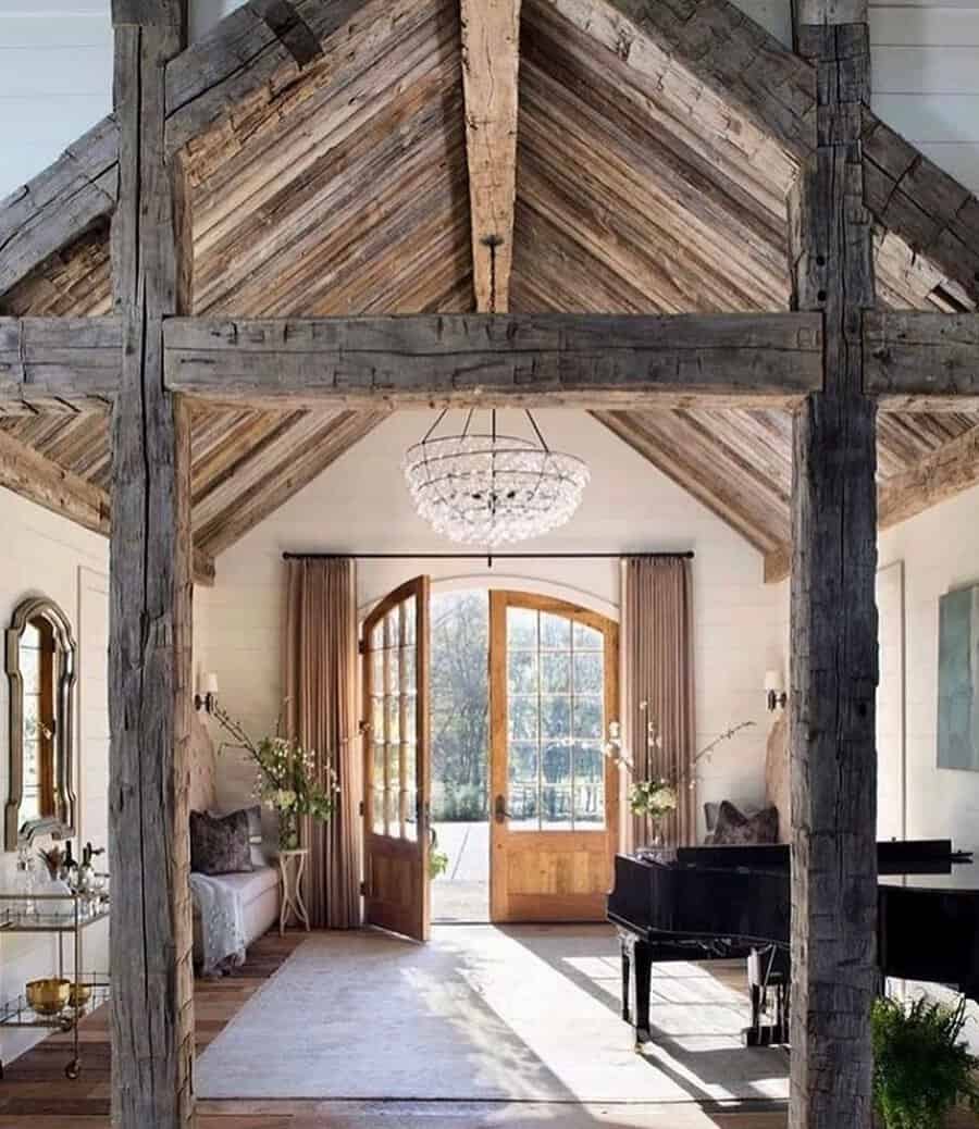 Vaulted space with reclaimed wood beams, a grand chandelier, and large wooden doors, framing a piano and airy seating area.