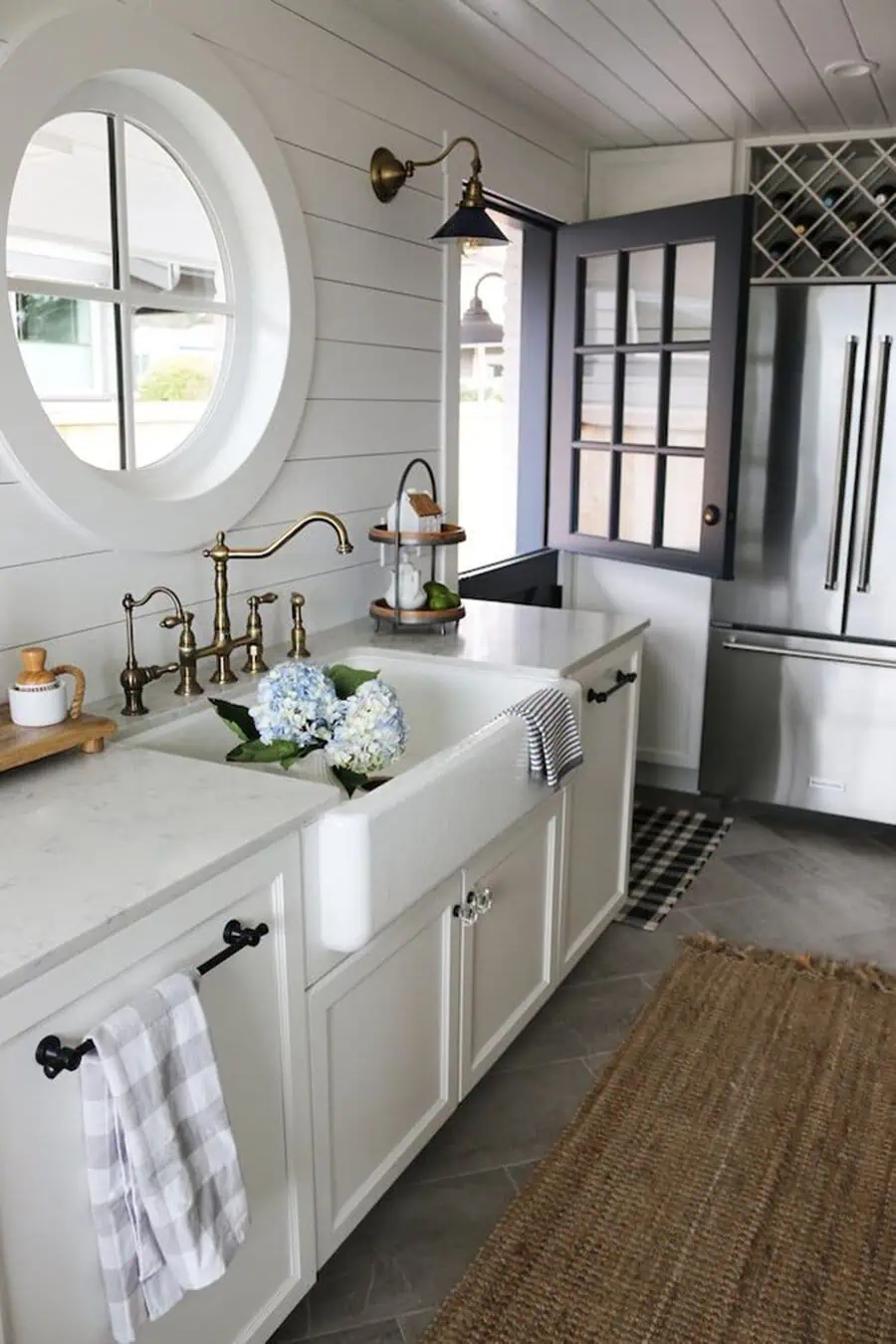 White kitchen with round window, farmhouse sink, brass fixtures, and shiplap walls, creating a charming coastal feel.
