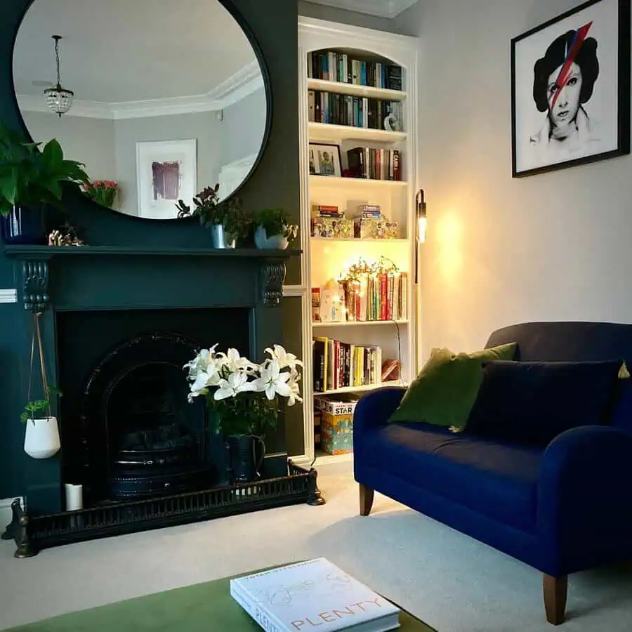 Large round mirror above a fireplace with lilies, navy sofa, and a bookshelf illuminated with string lights in a cozy living room.