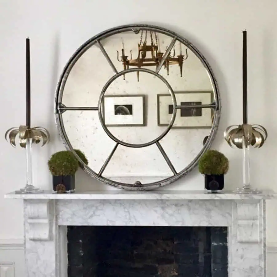 Antique-style round mirror on a marble fireplace, reflecting a vintage chandelier, with moss plants and sculptural candle holders for a classic look.