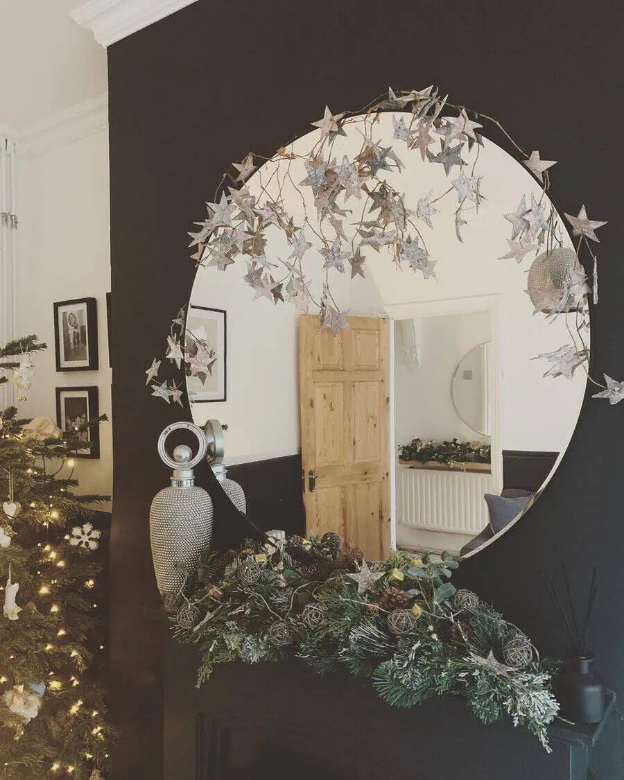 Round mirror framed with a star garland above a festive mantel with greenery, pinecones, and a Christmas tree in a cozy living room.