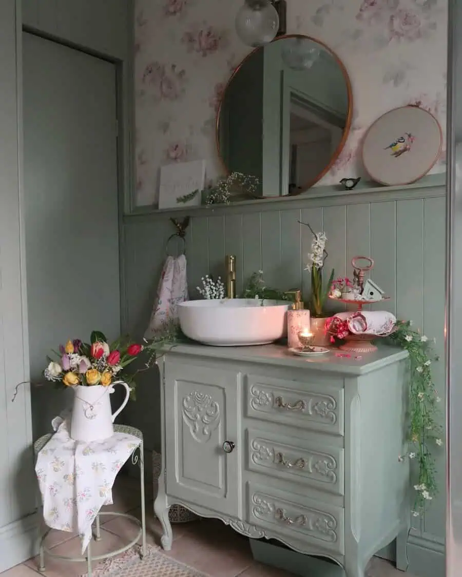 Vintage-style bathroom with a round mirror, mint green vanity, vessel sink, flowers, and candles, complemented by floral wallpaper and decor