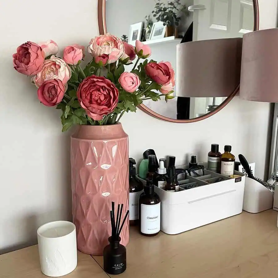 Round pink-framed mirror above a table with pink flowers in a vase, neatly organized beauty products, and candles.
