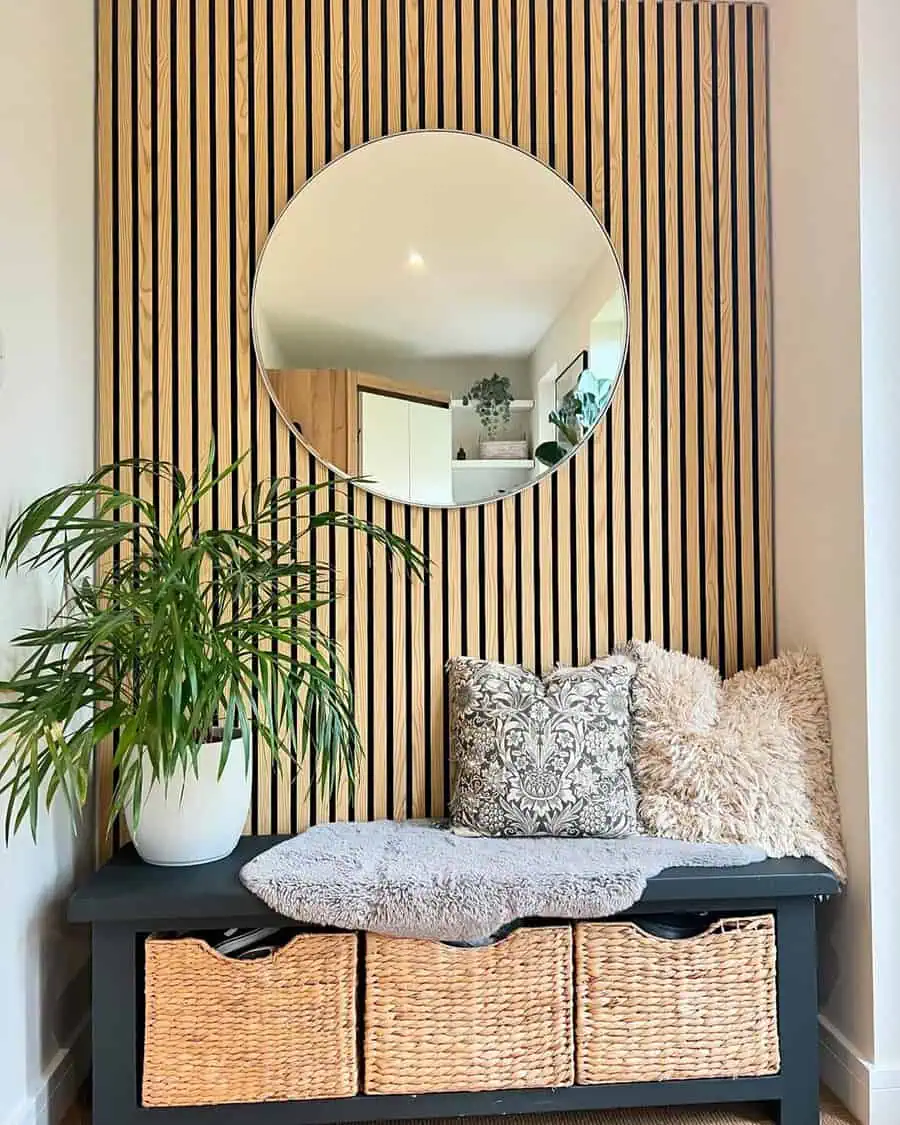Entryway with a round mirror on a slatted wood wall, a dark bench with woven baskets, cozy cushions, and lush green plants.