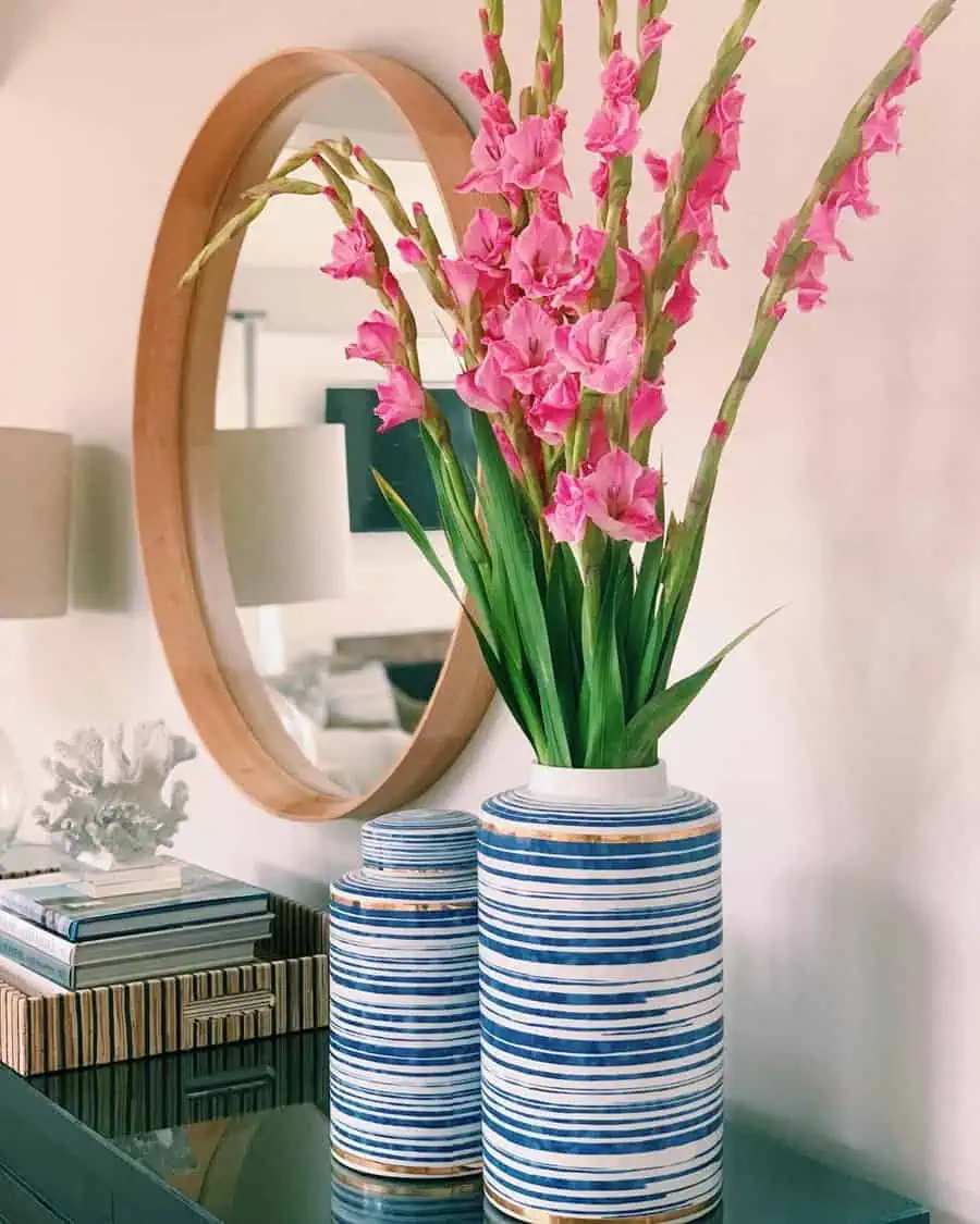 Round mirror with pink gladiolus flowers and striped blue vases on a console table with books and accessories.