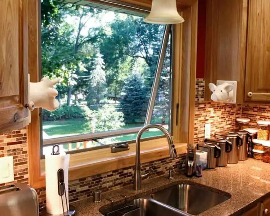 Wood-framed awning window in a warm kitchen with mosaic tile and a lush green view.