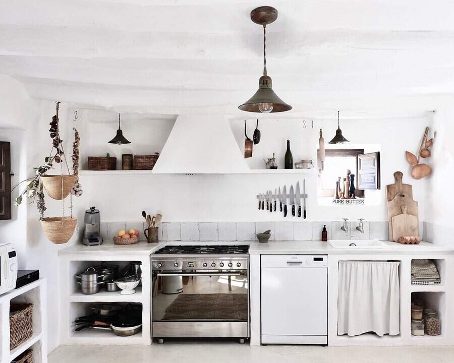 Rustic Spanish kitchen with white plaster walls, built-in shelves, woven baskets, and a linen curtain under the sink.
