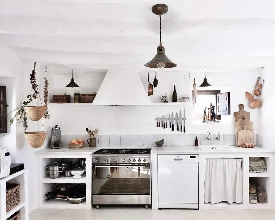 Rustic Spanish kitchen with white plaster walls, built-in shelves, woven baskets, and a linen curtain under the sink.