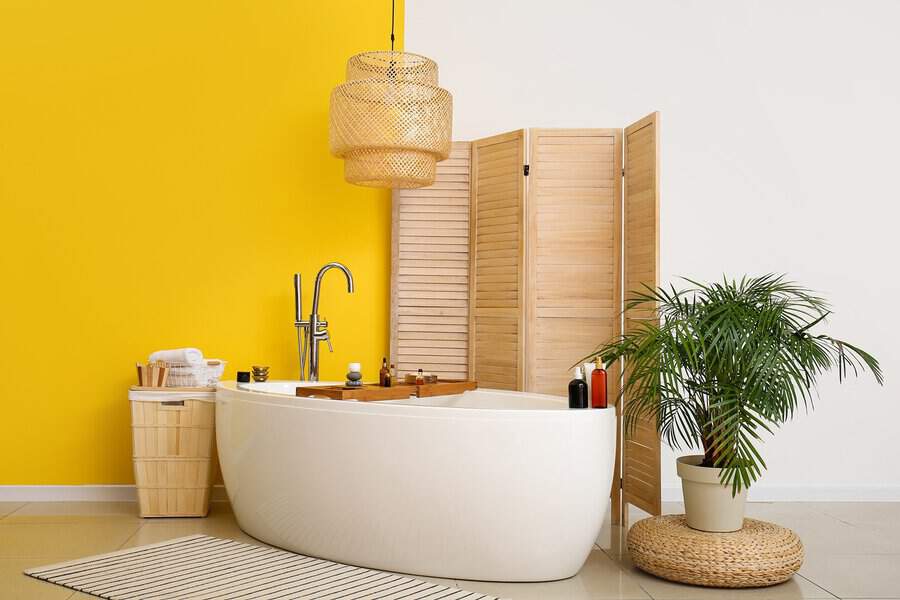 Freestanding white bathtub against a bold yellow wall with wood accents, a rattan pendant light, and a tropical plant for a spa-like feel.