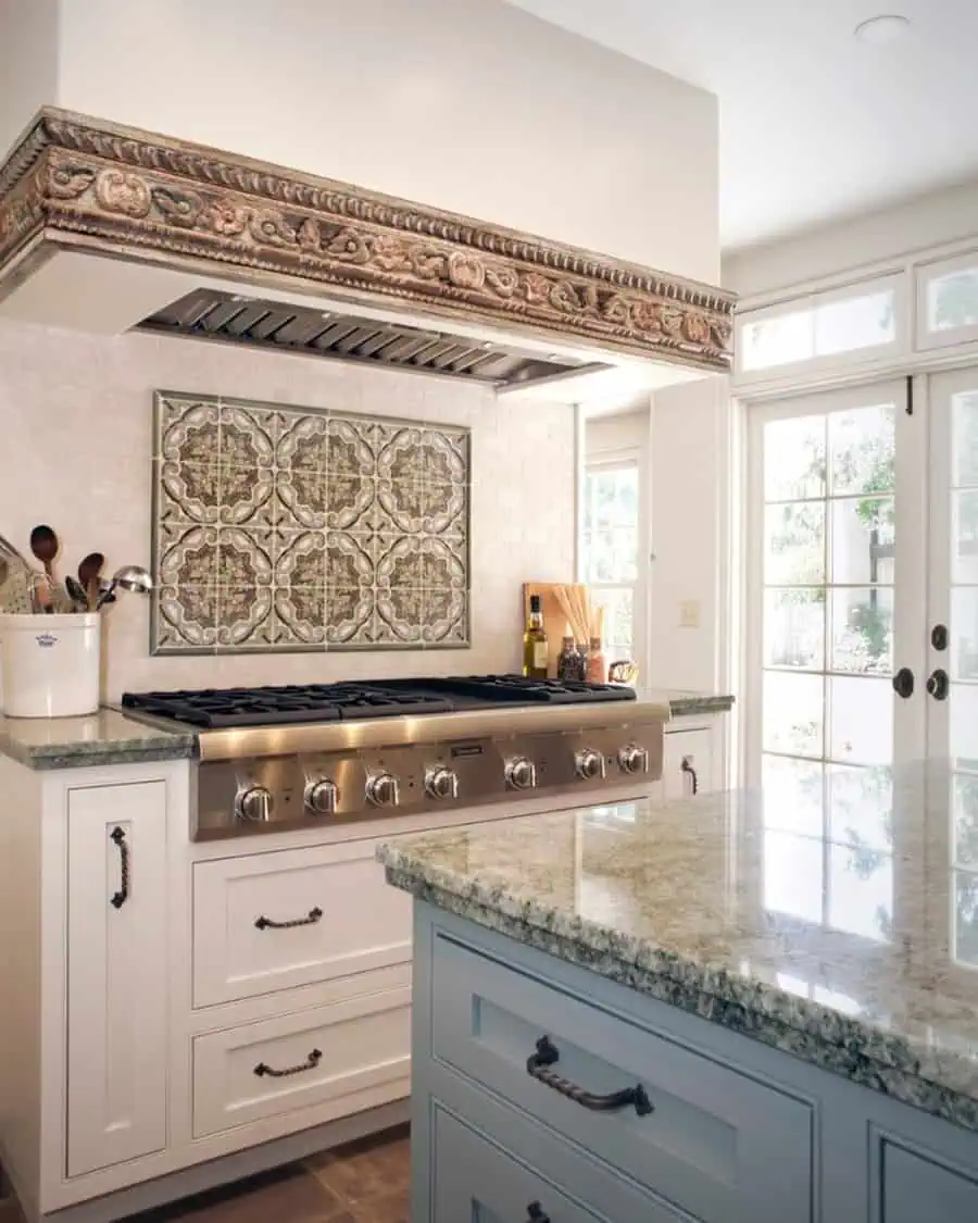 Spanish-style kitchen with ornate tile backsplash, carved wood range hood, a blue island, and natural stone countertops.