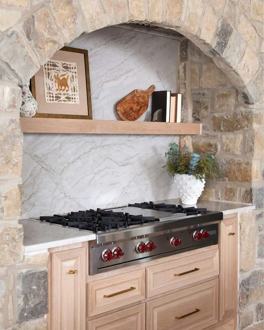 Spanish-style kitchen with a stone arch over the range, light wood cabinetry, a marble backsplash, and a floating shelf with decor.