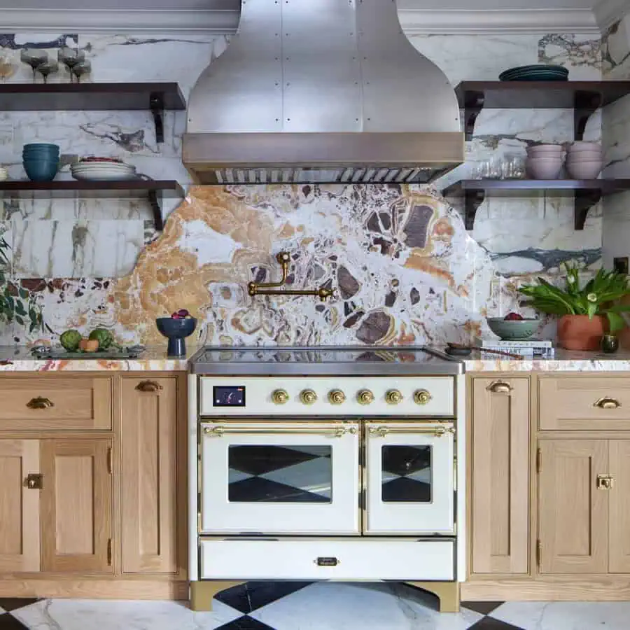 Spanish-style kitchen with a bold stone backsplash, brass fixtures, light wood cabinetry, open shelving, and a white and gold range.