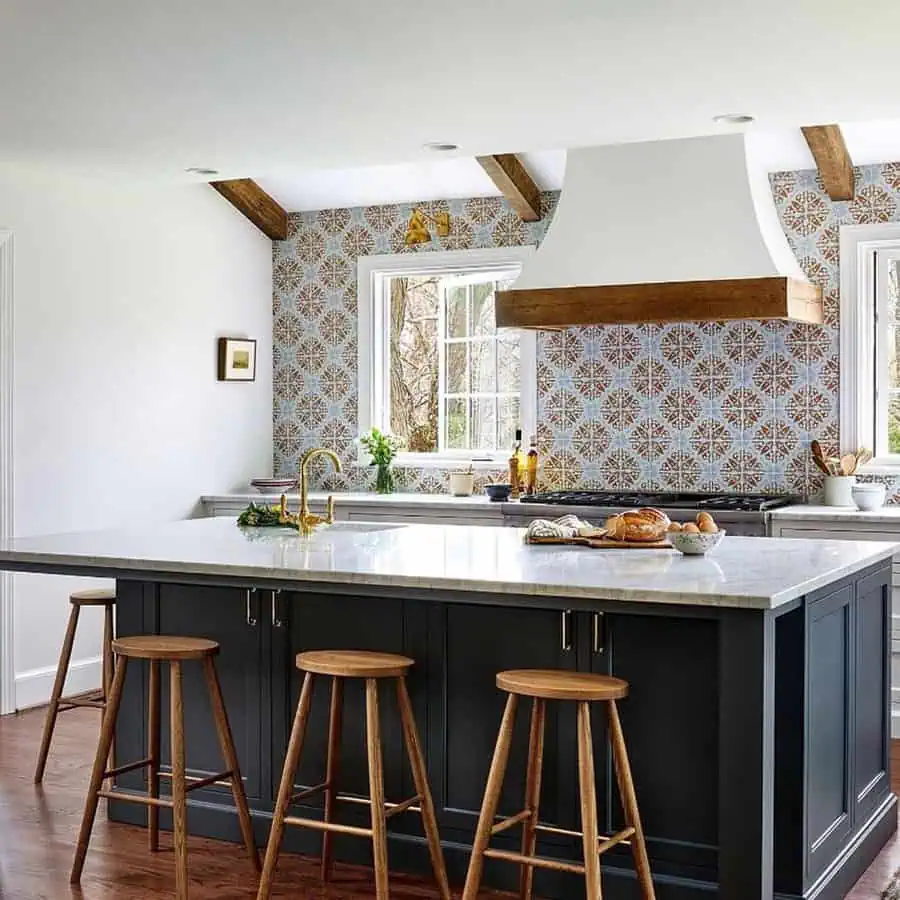 Spanish-style kitchen with an encaustic tile backsplash, wood beams, a white range hood, brass fixtures, and a dark blue island with wooden stools.