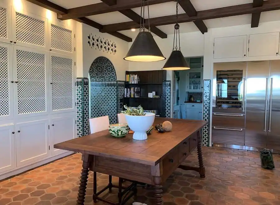 Spanish-style kitchen with patterned tile, terracotta flooring, exposed wood beams, lattice cabinets, and a rustic wooden dining table.