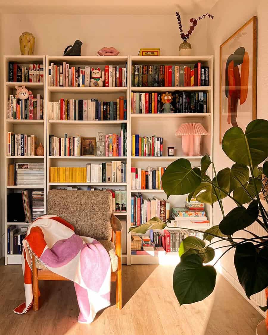 Sunlit corner with white bookshelves, a mid-century chair, colorful throw, and a large houseplant for a cozy reading nook.