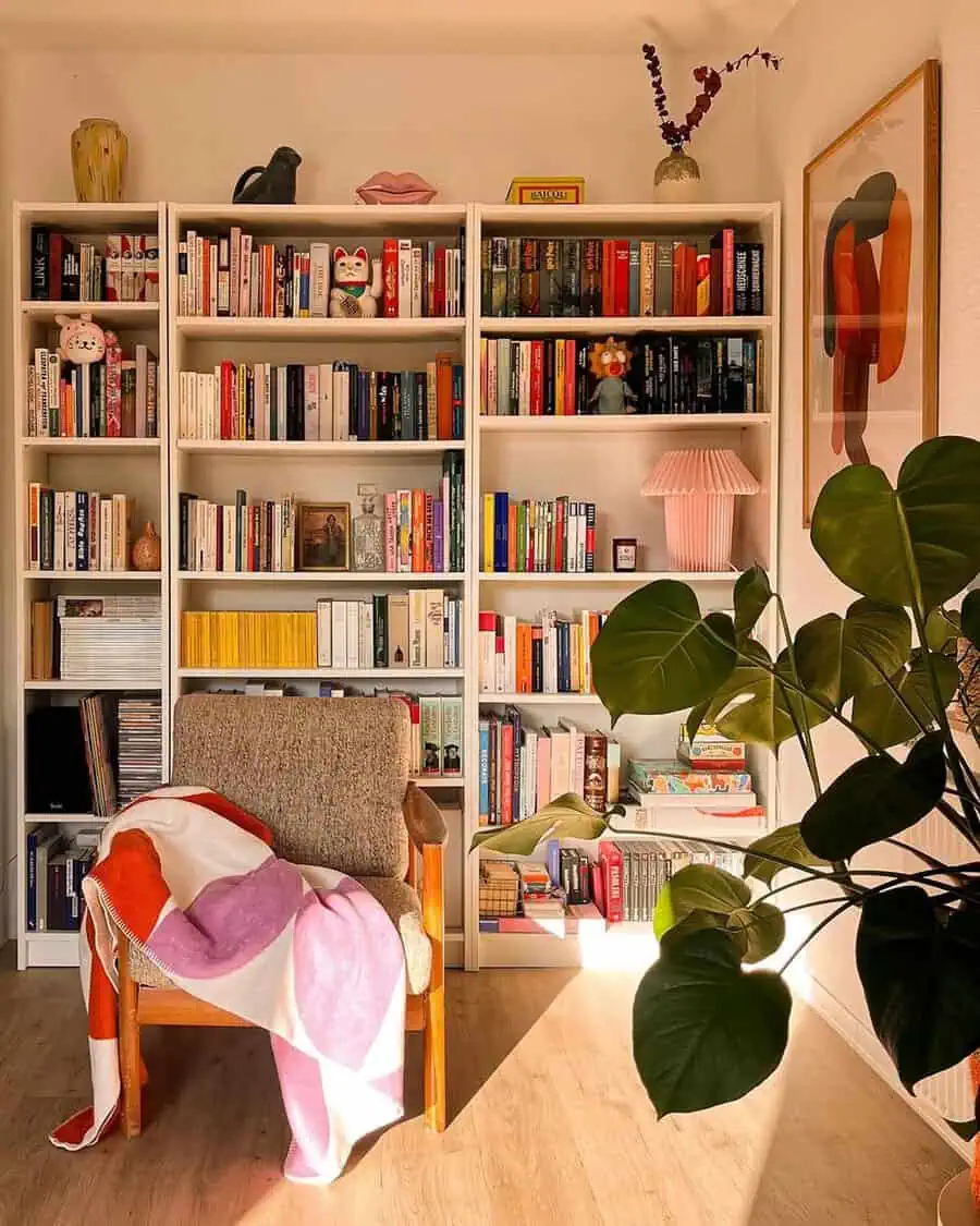 Sunlit corner with white bookshelves, a mid-century chair, colorful throw, and a large houseplant for a cozy reading nook.