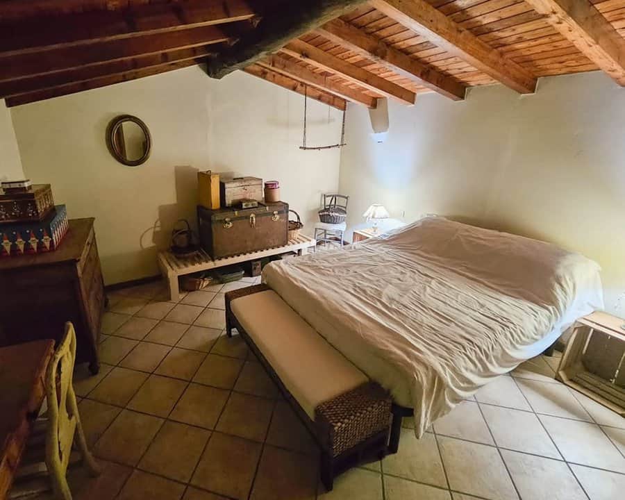 Attic spare room with wooden beams, antique trunks, neutral bedding, and a cozy bench for a rustic, vintage-inspired retreat.