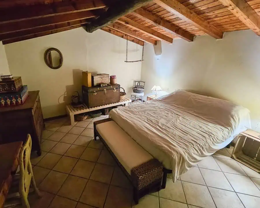 Attic spare room with wooden beams, antique trunks, neutral bedding, and a cozy bench for a rustic, vintage-inspired retreat.