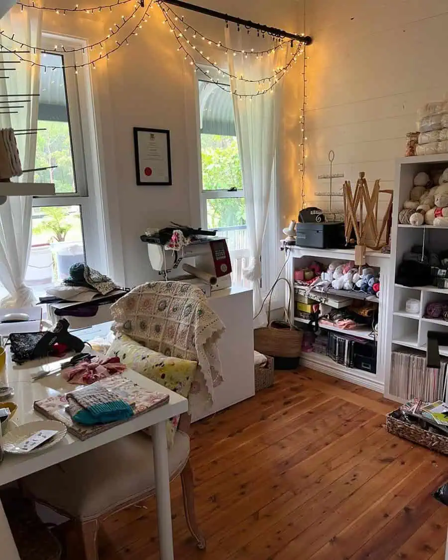 Craft room with string lights, shelves of yarn, sewing machine, and sunlight through sheer curtains for a warm, creative nook.