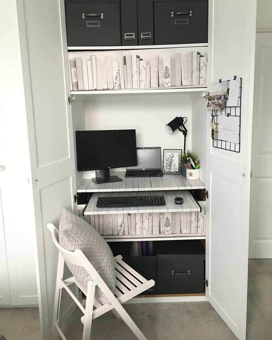 Wardrobe office with pull-out shelves, black storage boxes, book-patterned wallpaper, a folding chair, and a mounted desk lamp.