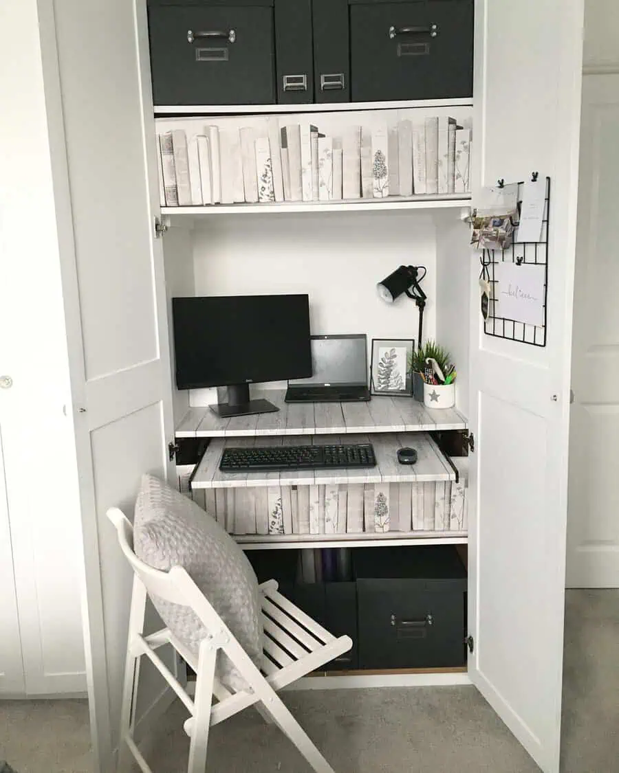 Wardrobe office with pull-out shelves, black storage boxes, book-patterned wallpaper, a folding chair, and a mounted desk lamp.