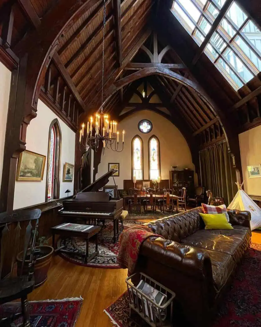 Converted church living space with high wooden beams, stained glass windows, chandelier, grand piano, and leather sofa.
