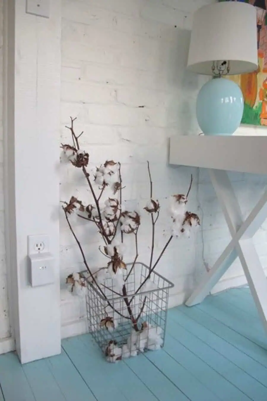Turquoise-painted wood floor with white brick walls, a small desk, and cotton stems in a wire basket.